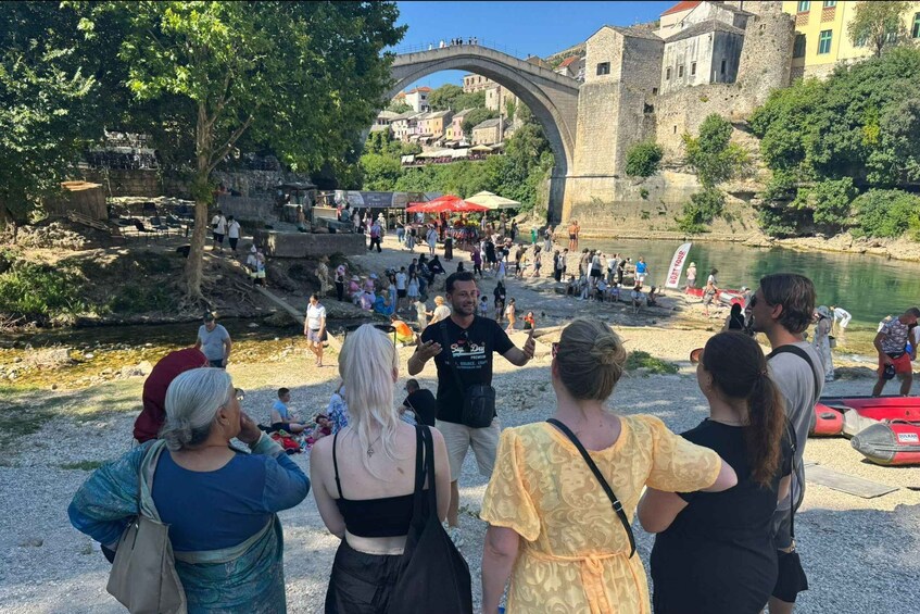 A group of people standing by a river, a stone bridge, and historic buildings in the background. 1