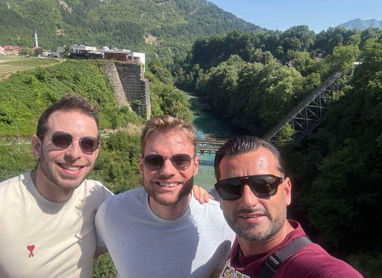 Three people standing on a bridge with a river and mountains in the background. 3