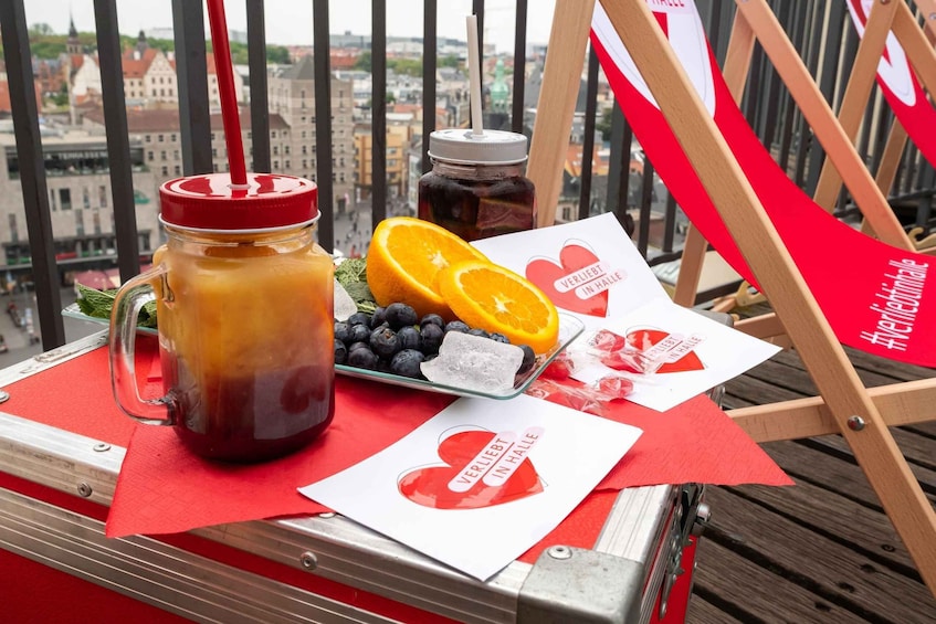 A balcony with a red table holding a glass jar of iced tea, a plate of blueberries, and an orange slice. There are also two cards with the text "VERLIET IN HALLE" and a hashtag "#VERLIETINHALLE". 4
