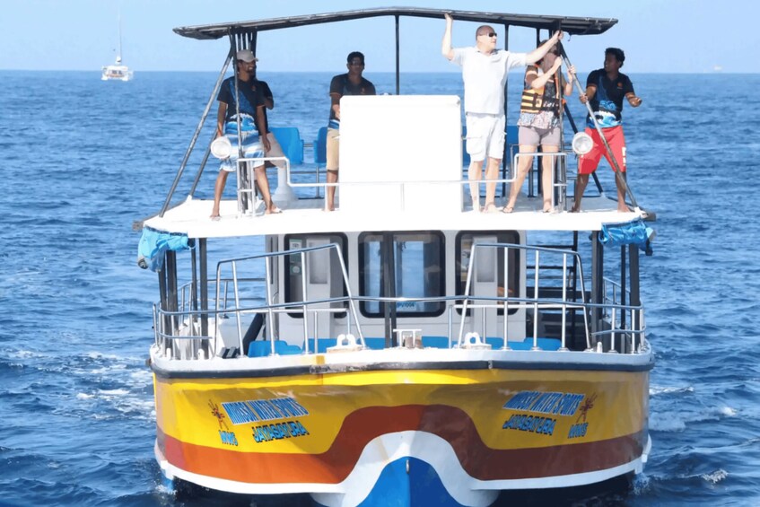A colorful boat with people standing on the deck, a clear blue sea, and a distant sailboat. 4