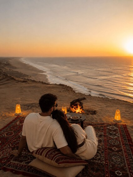 A couple sitting on a patterned rug by a beach at sunset, with a fire between them. 3