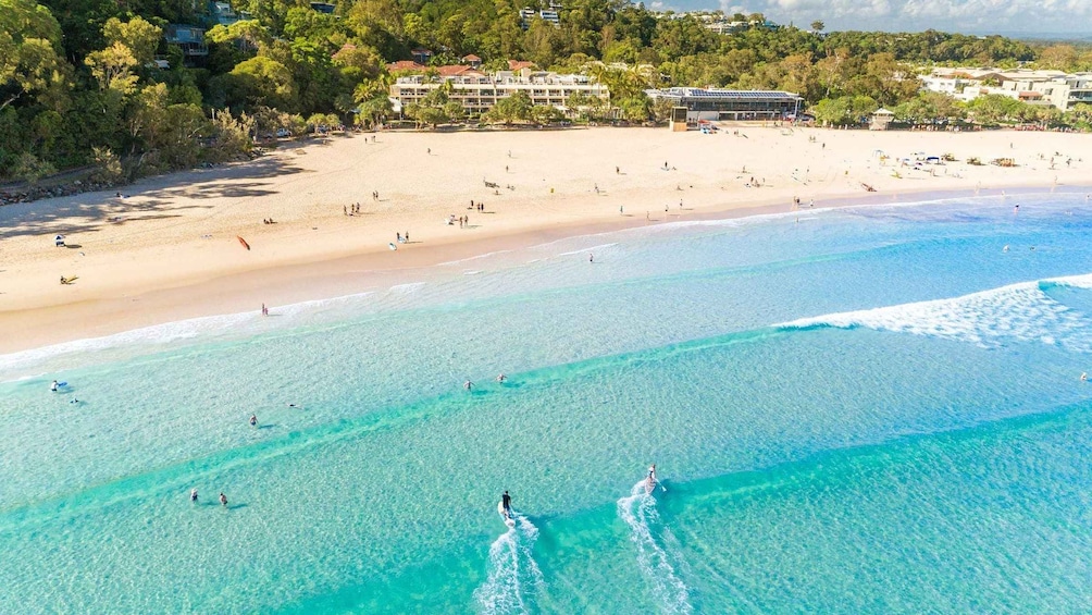A beach with clear turquoise water, people swimming and surfing, and buildings in the background. 4