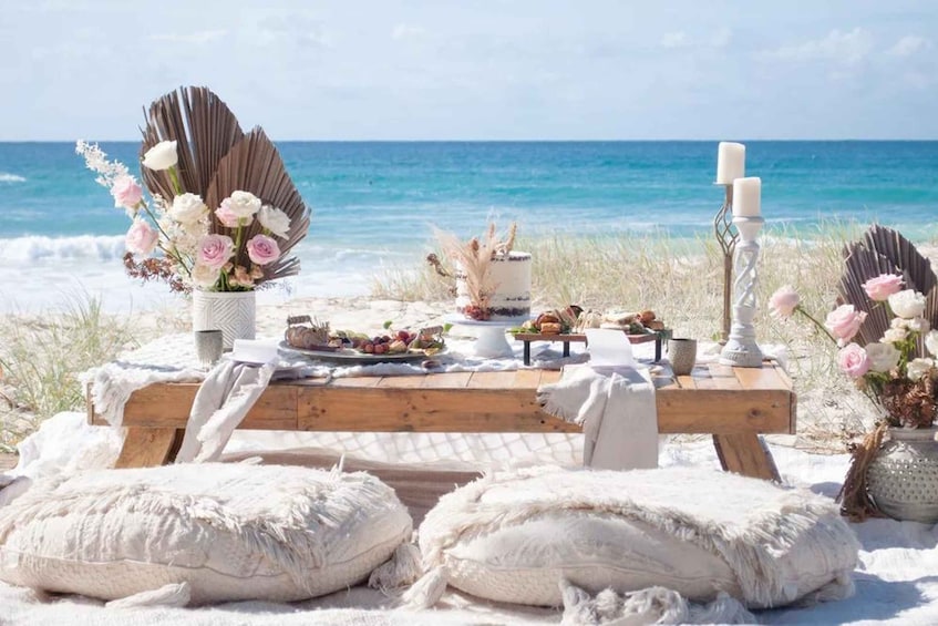 A beach picnic setup with a wooden table, white cushions, a cake, flowers, and a candle. 3