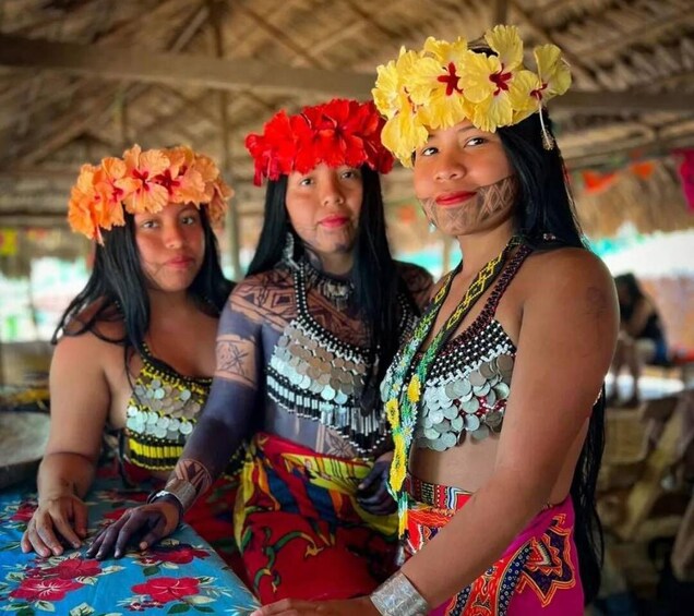 Drei Personen in traditioneller Kleidung mit floralen Kopfschmuck und Perlenketten. 1