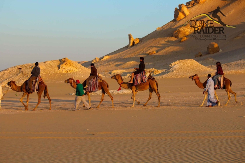 A group of people on camels in a desert landscape with sand dunes and rock formations. 1