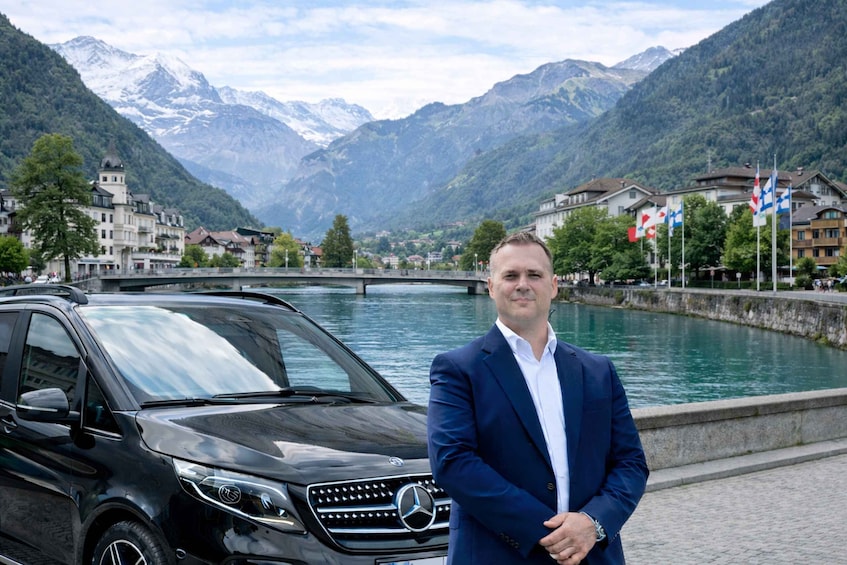 A man in a blue suit standing next to a black Mercedes-Benz SUV by a river with mountains in the background. 5