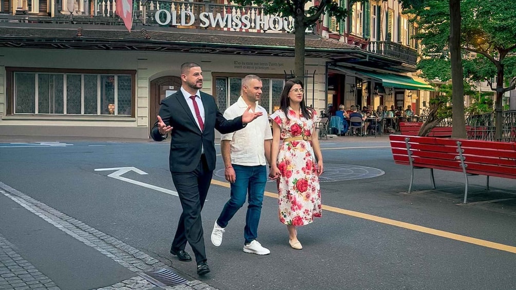A man in a suit gesturing, a woman in a floral dress, and a man in a white shirt walking on a street with a building labeled 'OLD SWISS HOUSE' in the background. 2