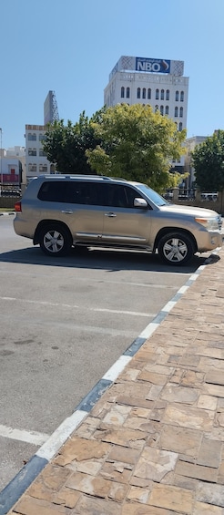 A beige SUV parked on a city street with a stone-paved sidewalk. In the background, there is a building with a sign that reads "NBO". 5