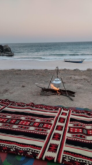 A beach with a fire, a boat, and a patterned blanket. 2