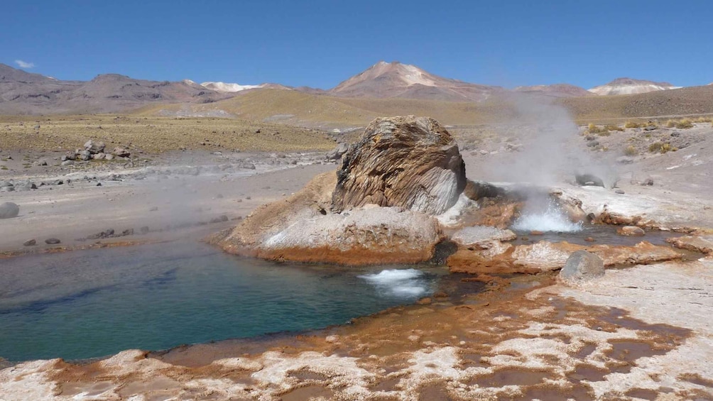 A geothermal area with a hot spring, a rocky outcrop, and a mountainous background. 2