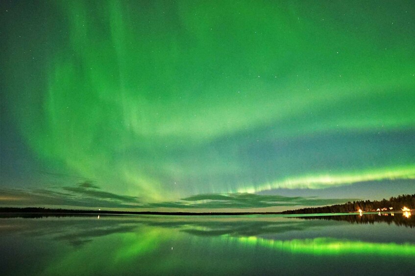 The Northern Lights over a calm lake with reflections, a shoreline, and distant lights. 3