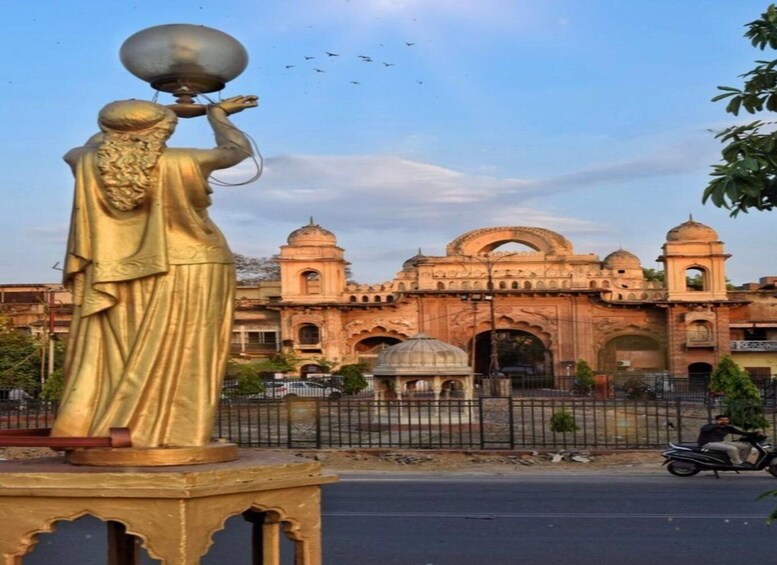 A golden statue of a figure holding a pot, in front of a historic building with arches and domes. 2