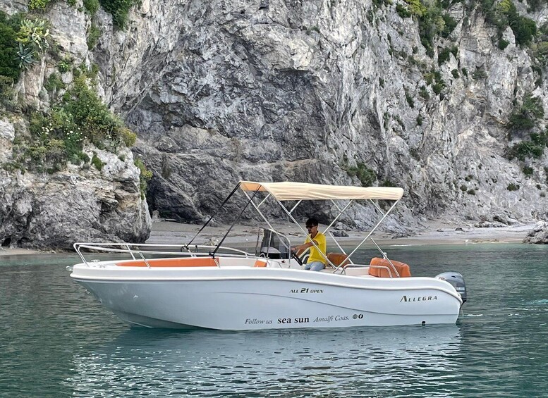 En hvid båd, der hedder 'Amalfi Coasts', med en person i en gul skjorte om bord, tæt på klipper. 3