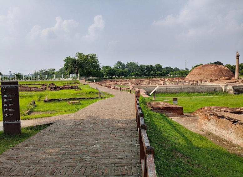A paved pathway leading through an archaeological site with brick ruins and a large mound in the background. 2