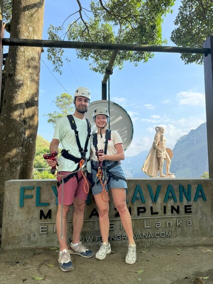Two people in harnesses standing in front of a sign that reads 'FLYAVANA MOUNTAIN LINE' with a statue of a figure in the background. 1