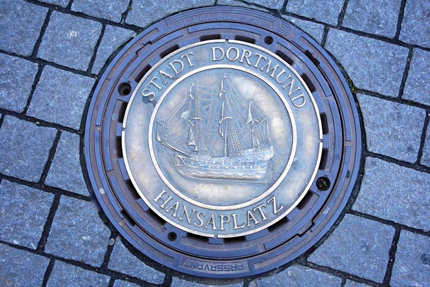 A manhole cover with the inscription "STADT DORTMUND HANSAPLATZ" and a ship engraving. 3
