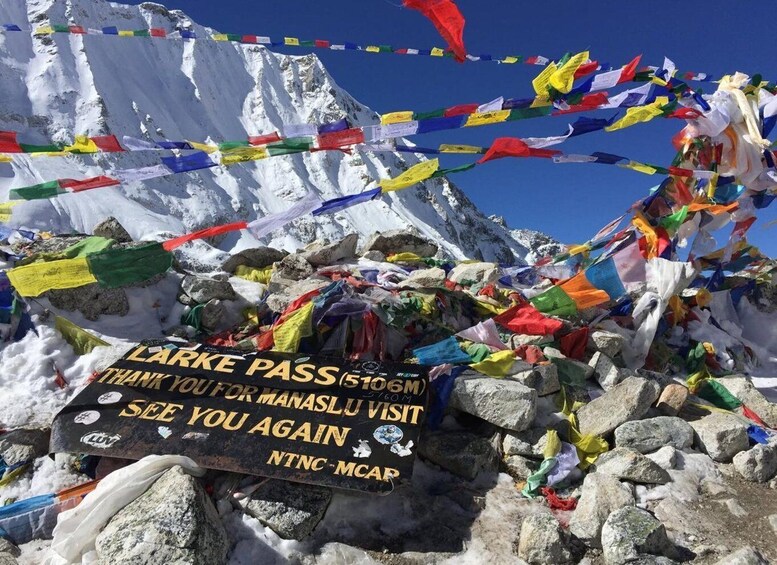 A mountain peak with colorful prayer flags and a sign thanking visitors for their support. 3