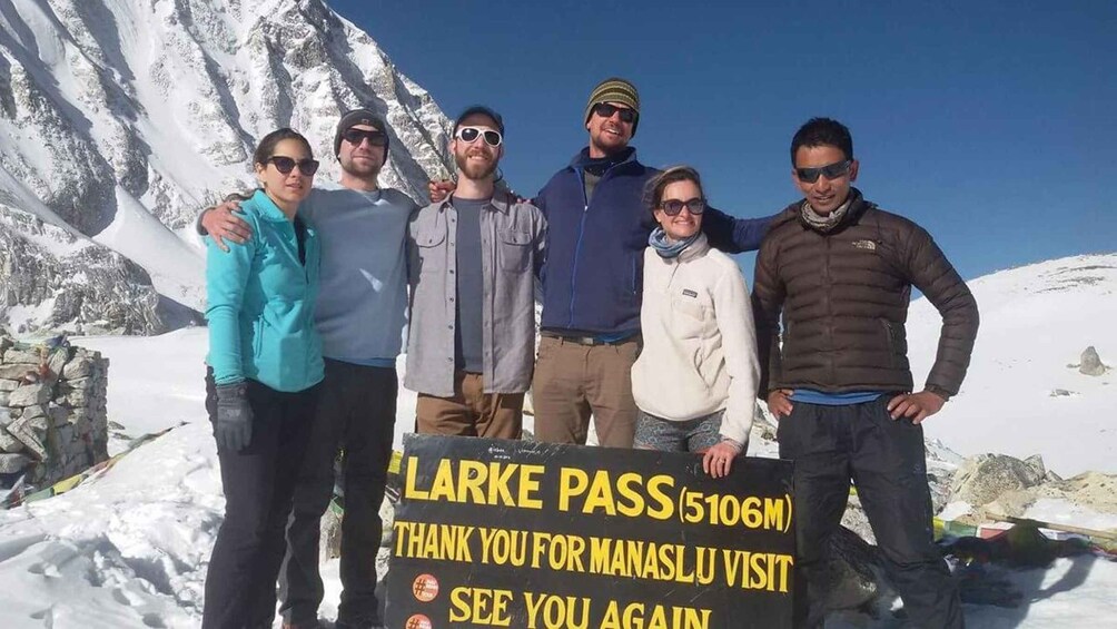 A group of people standing in a snowy mountainous area with a sign that reads "LARKE PASS (5106M) THANK YOU FOR MANASLU VISIT SEE YOU AGAIN". 1