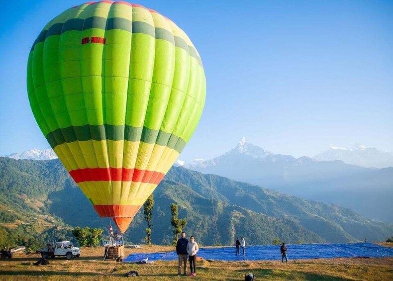 A hot air balloon with a green and yellow color scheme, a vehicle, and a mountainous landscape in the background. 1