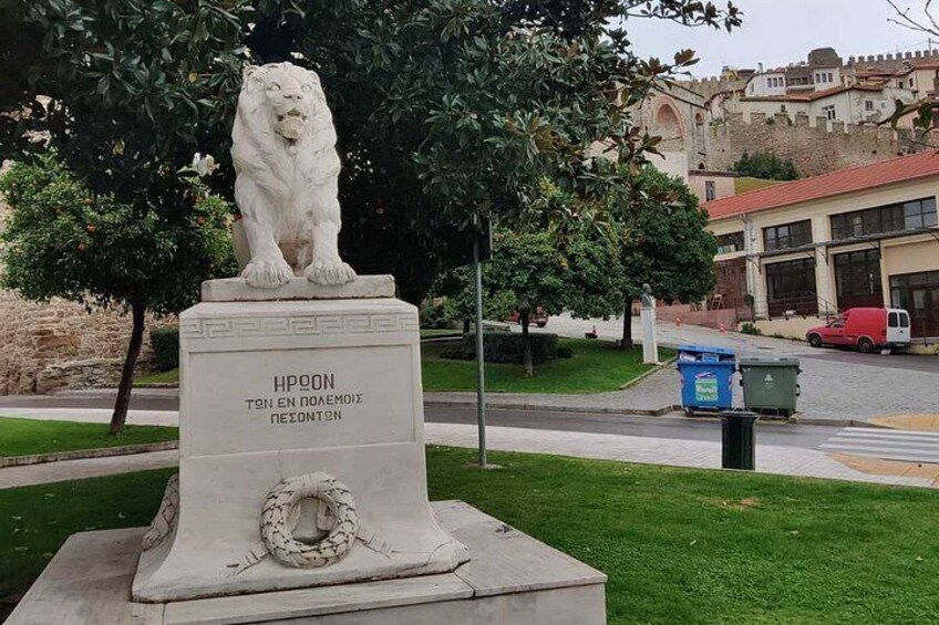 A white lion statue on a pedestal with a wreath, surrounded by grass and trees, with a building and a red vehicle in the background. 4