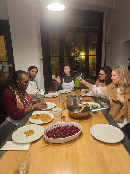 A group of people dining at a wooden table with plates of food, a bowl of salad, and a pot on the table. 5