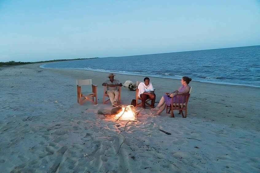 Three people sitting by a campfire on a beach with the ocean in the background. 1