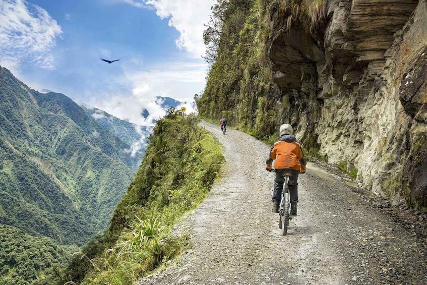 A mountain biker on a narrow, rocky path with steep cliffs on one side and a lush valley below. 1