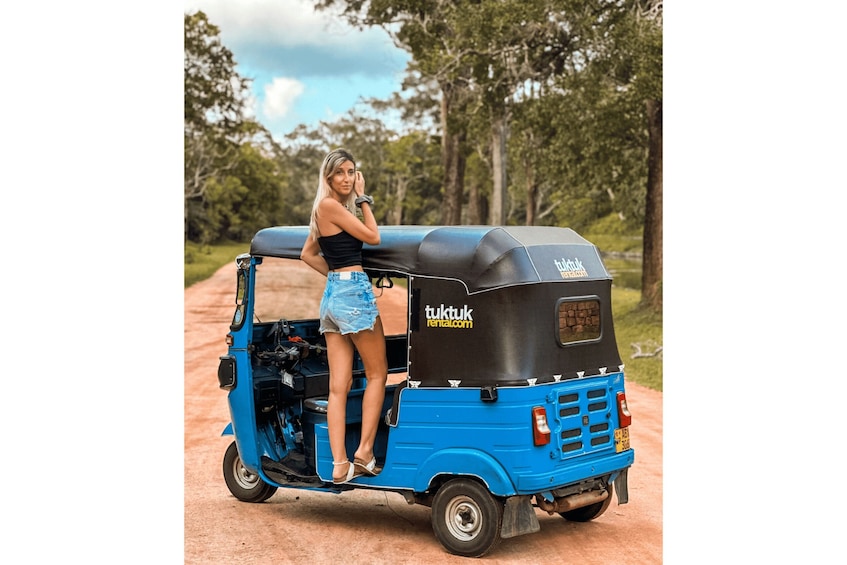 A woman sitting on a blue auto-rickshaw parked on a dirt road with trees in the background. 5