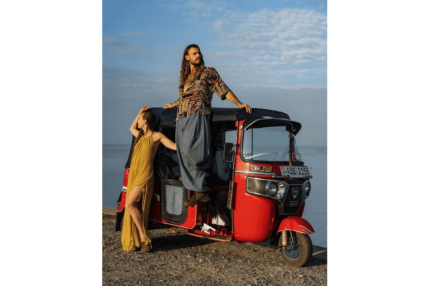 A red auto-rickshaw parked on a rocky shore with a person leaning on it and another person sitting inside. 3