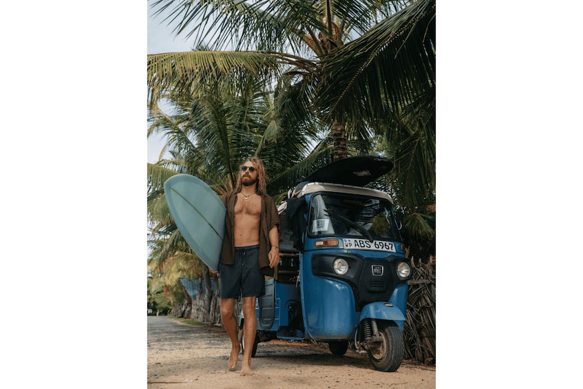 A man with a surfboard standing next to a blue auto-rickshaw. 4