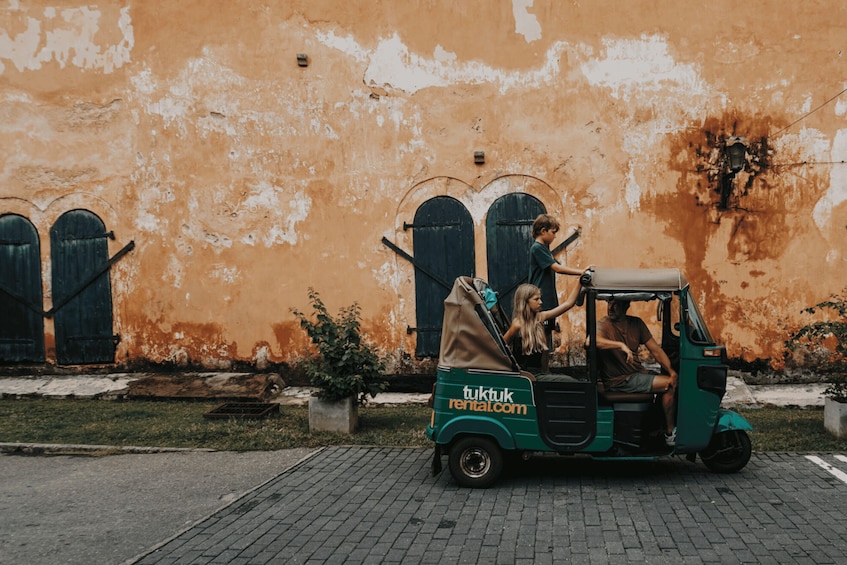 A tuk-tuk with two passengers, a man and a woman, parked in front of a weathered orange wall with arched windows. 2