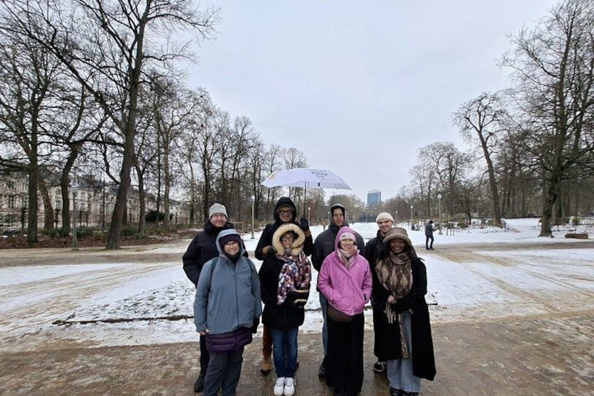 A group of people standing on a snow-covered path in a park with leafless trees and buildings in the background. 2