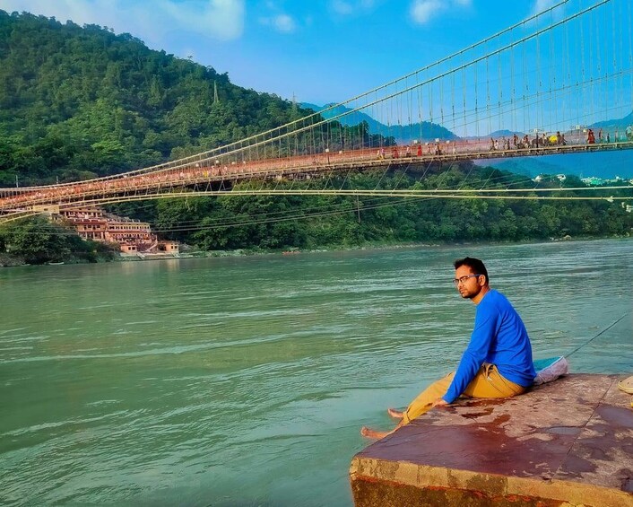 A man sitting by a river, a bridge in the background, and a forested hillside. 3