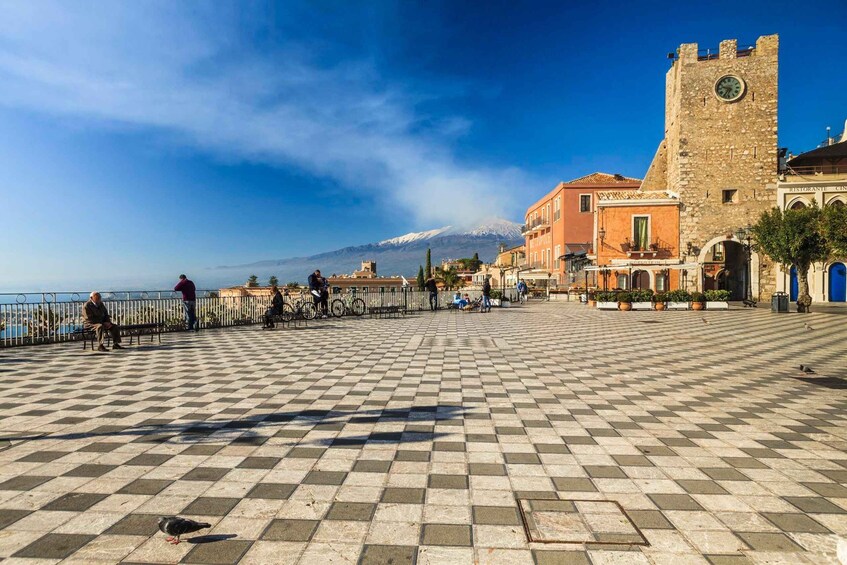A checkered plaza with a historic tower, a café, and a view of a mountain. 1