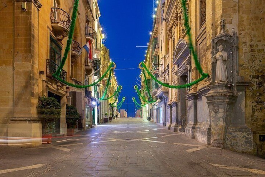 Calle iluminada de noche, flanqueada por edificios decorados con guirnaldas verdes. 3