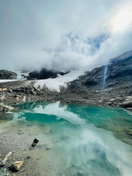 A glacier with a clear turquoise pool at its base, surrounded by rocky terrain and misty clouds. 3