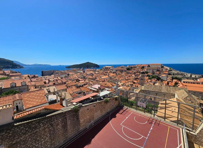 A coastal cityscape with terracotta-roofed buildings, a basketball court, and a clear blue sky. 1