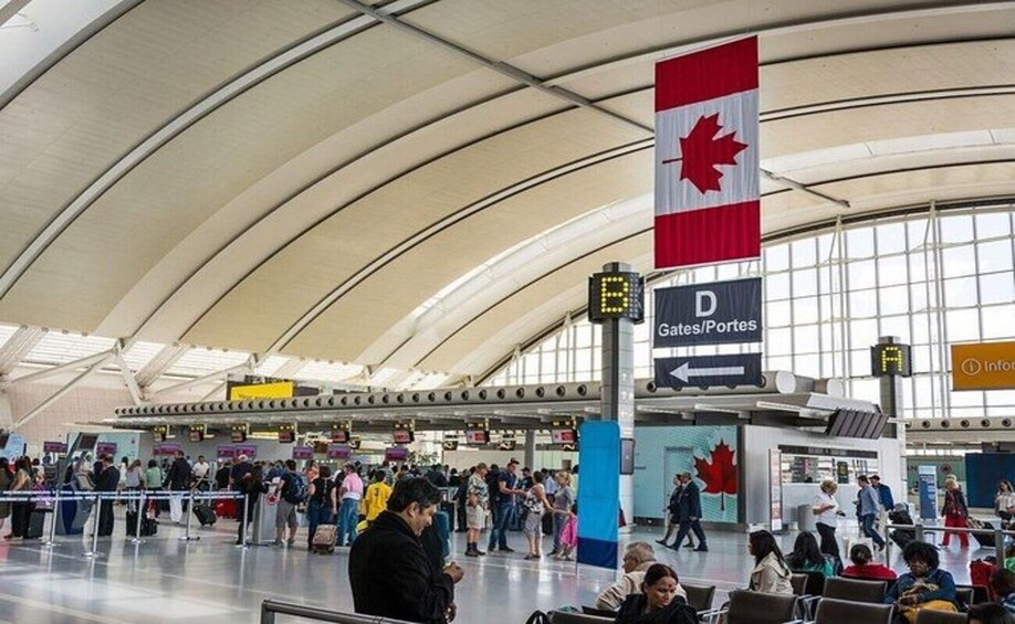 An airport terminal with a large Canadian flag, signs for Gates/Portes D, and a crowd of travelers. 2