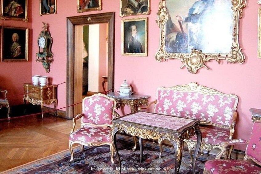 Een kamer met rijk ingericht meubilair, waaronder een roze bank en bijpassende stoelen, een tafel en een spiegel met een gedetailleerde lijst. 5