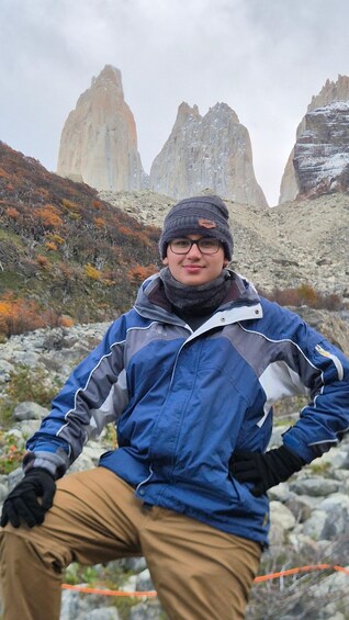 A person in a blue jacket and brown pants standing on rocky terrain with autumn-colored trees and mountain peaks in the background. 3