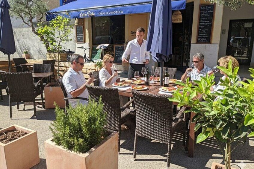 An outdoor cafe with wicker chairs and tables, a waiter serving food, and a blue awning. 2