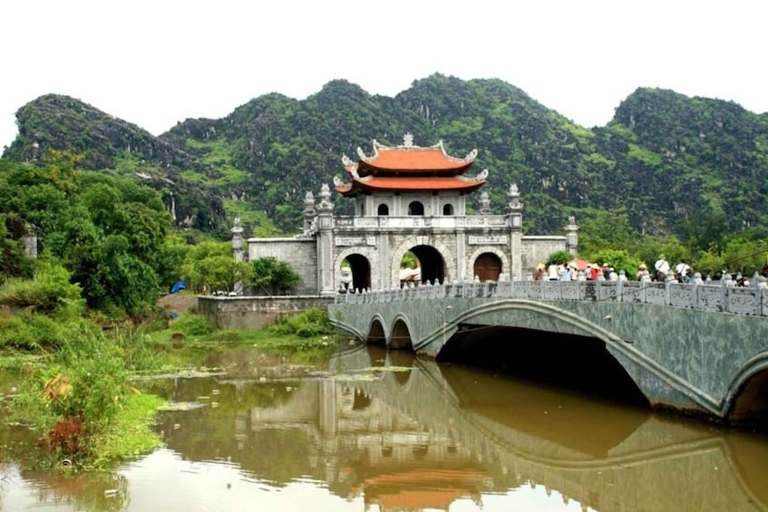 A traditional stone bridge with a red-tiled roof, leading to a gate with ornate carvings, surrounded by lush greenery and a river. 3