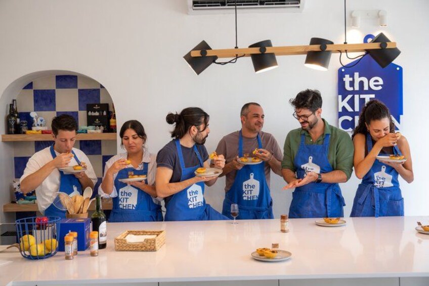 A group of people wearing aprons with 'the KIT CHEN' logo, standing around a table with plates of food. 2