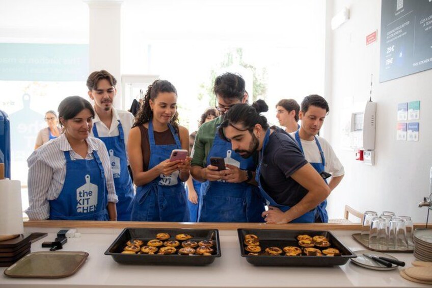 A group of people in aprons, some looking at a smartphone, in a kitchen setting with trays of food and kitchen equipment. 5