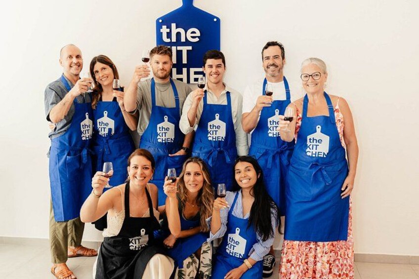 Un groupe de personnes portant des tabliers bleus ornés du logo « THE KIT CHEN », debout et assises devant une enseigne bleue portant le même logo. 1