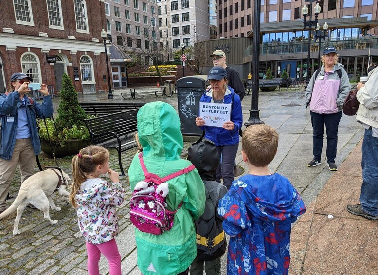 A group of people, including children, standing on a city street with a sign reading "BOSTON BY LITTLE FEET CHILDREN'S TOUR". 4