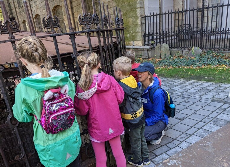 A group of children and an adult standing in front of a metal gate with a stone building in the background. 2