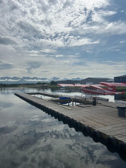 A calm lake with a dock, a red airplane with the registration N121KT, and mountains in the background. 4