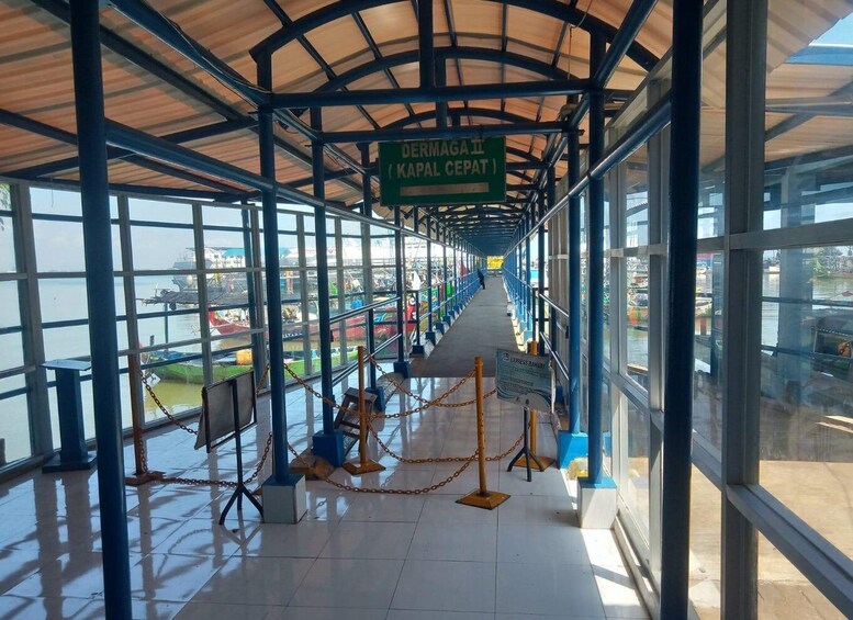 A covered walkway with a view of boats on water, a sign reading 'DERMAGA II (KAPAL DEPAT)', and a chain barrier. 5