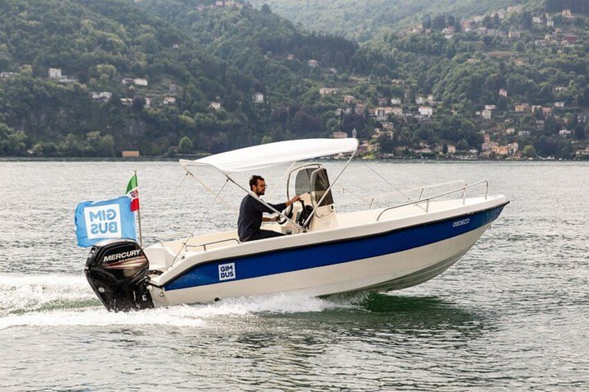 A motorboat with a white and blue color scheme, a person steering, and a backdrop of a lake with a hilly landscape. 1